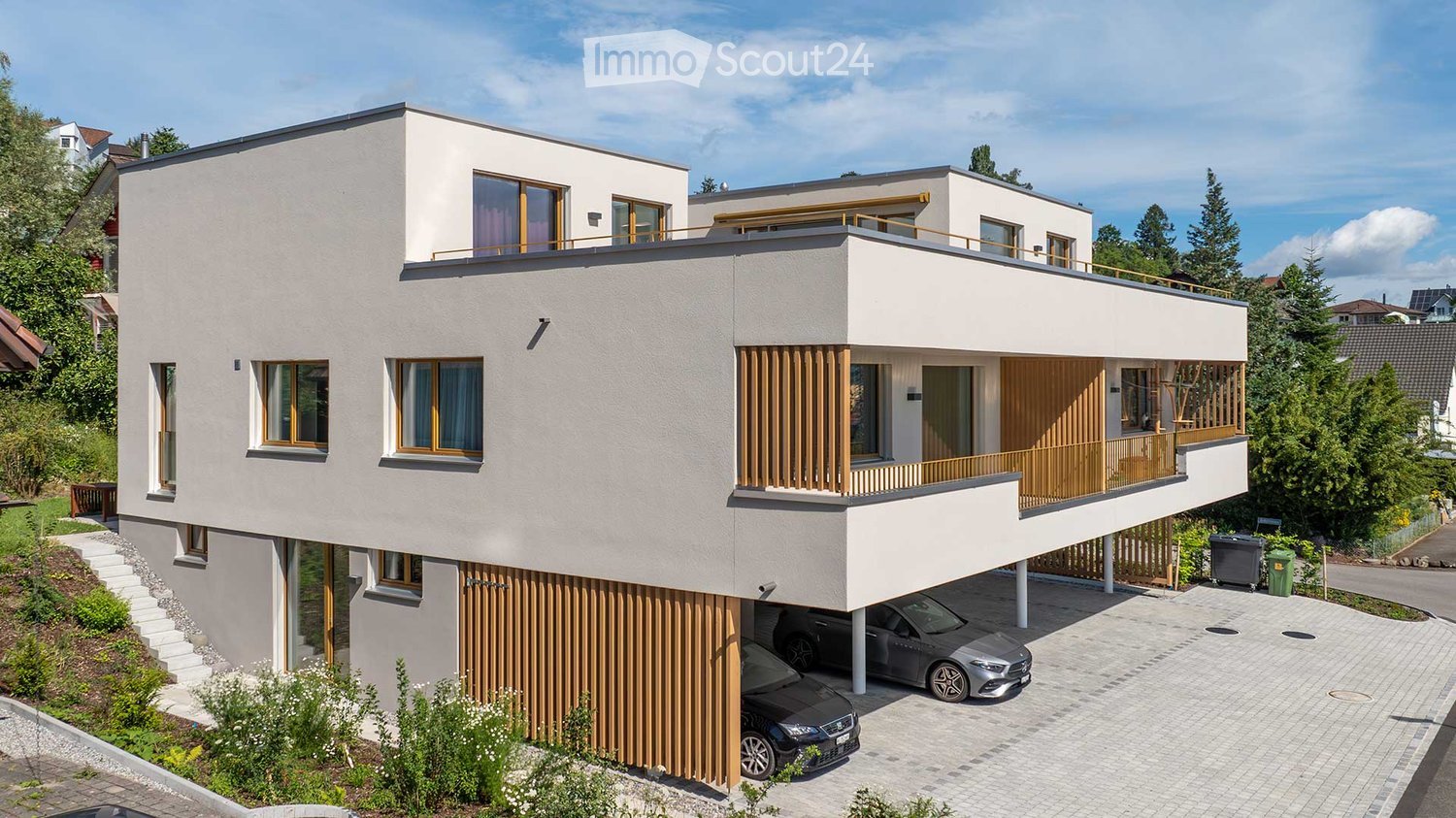 House. Modern two-story house with white facade, large windows, wooden shutters, and a balcony on the upper level. Garage with two cars parked inside, surrounded by greenery.