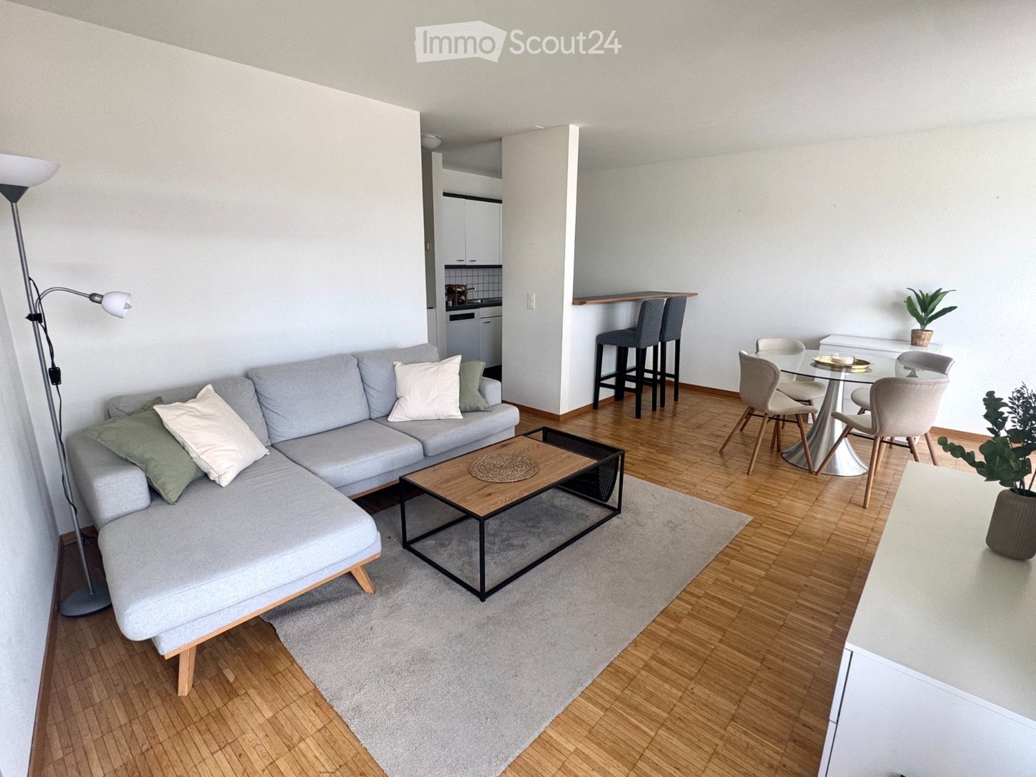 Modern living room with grey sectional sofa, wooden coffee table, dining area with glass table and chairs, and bar stools near the kitchen.