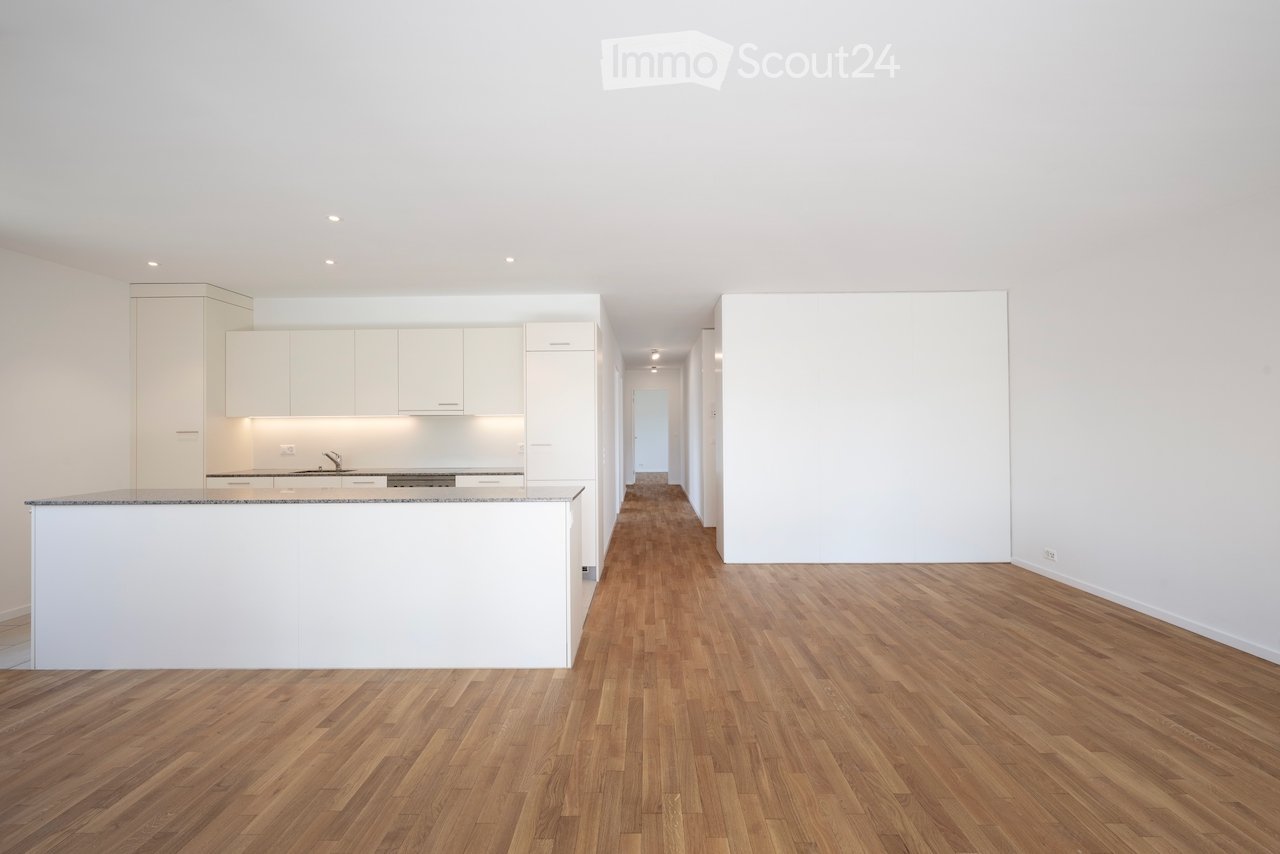 Open-plan unfurnished space with a modern kitchen area featuring white cabinets and a granite countertop, connected to a hallway with wooden flooring and white walls.