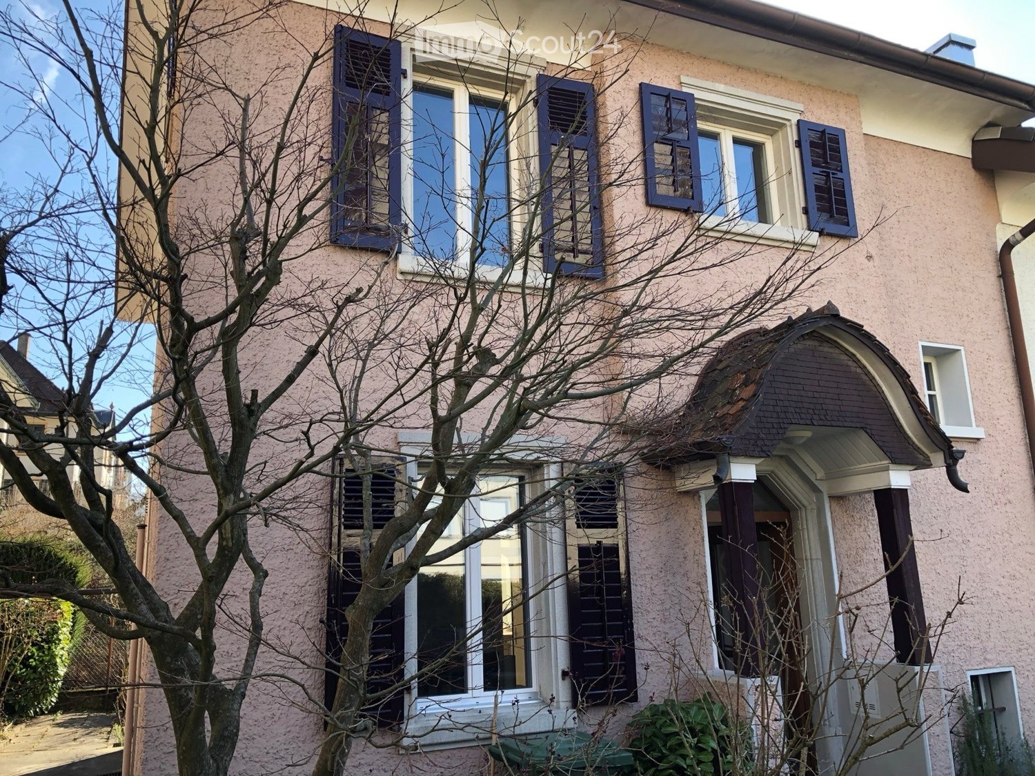 Two-story house with beige siding, multiple windows with shutters, and a decorative arched entrance.