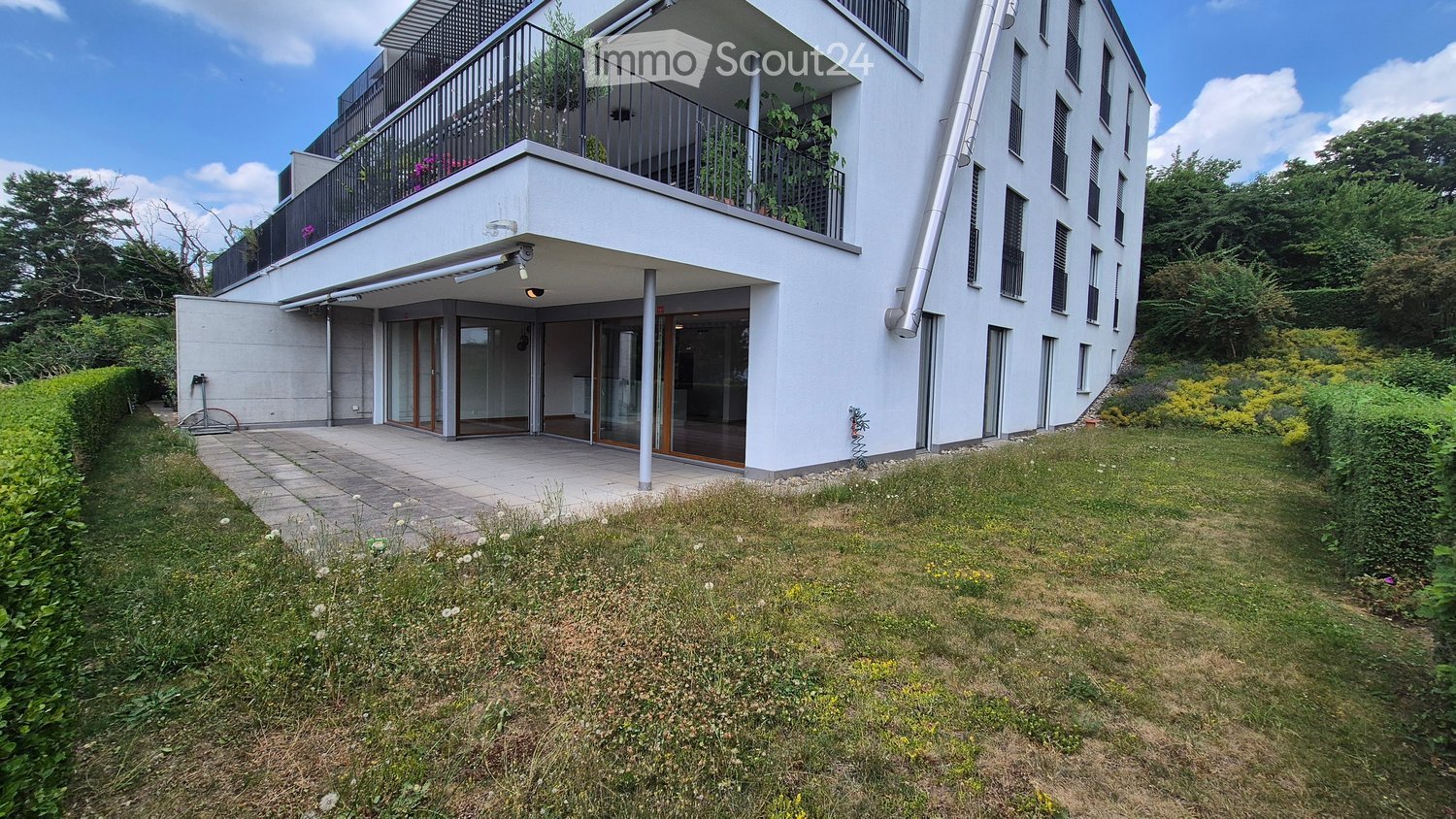 Modern white multi-story building with large windows, balcony, and surrounding greenery.