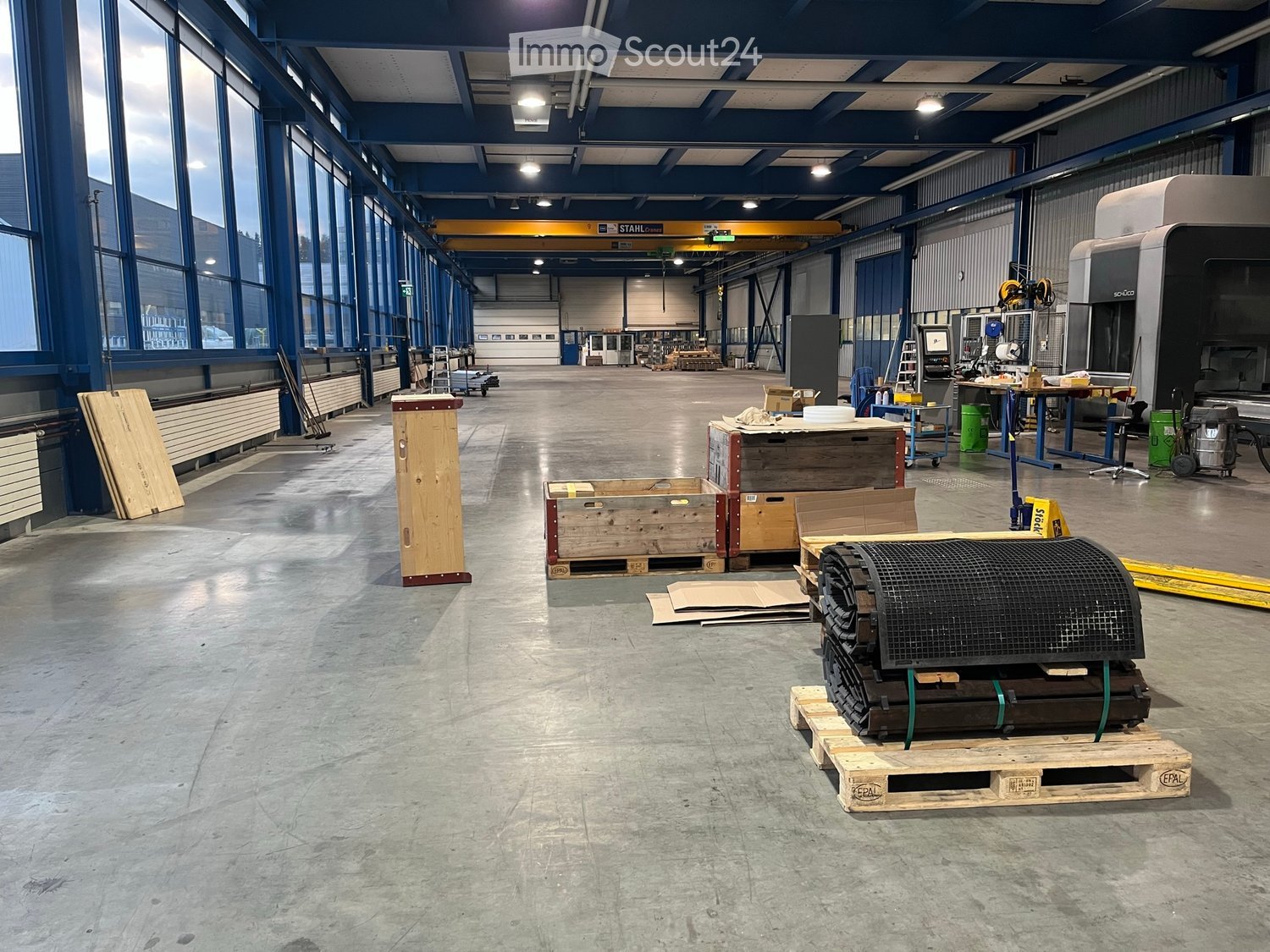 empty large factory space, blue metal pillars, grey flooring, storage boxes, large glass windows, yellow overhead crane