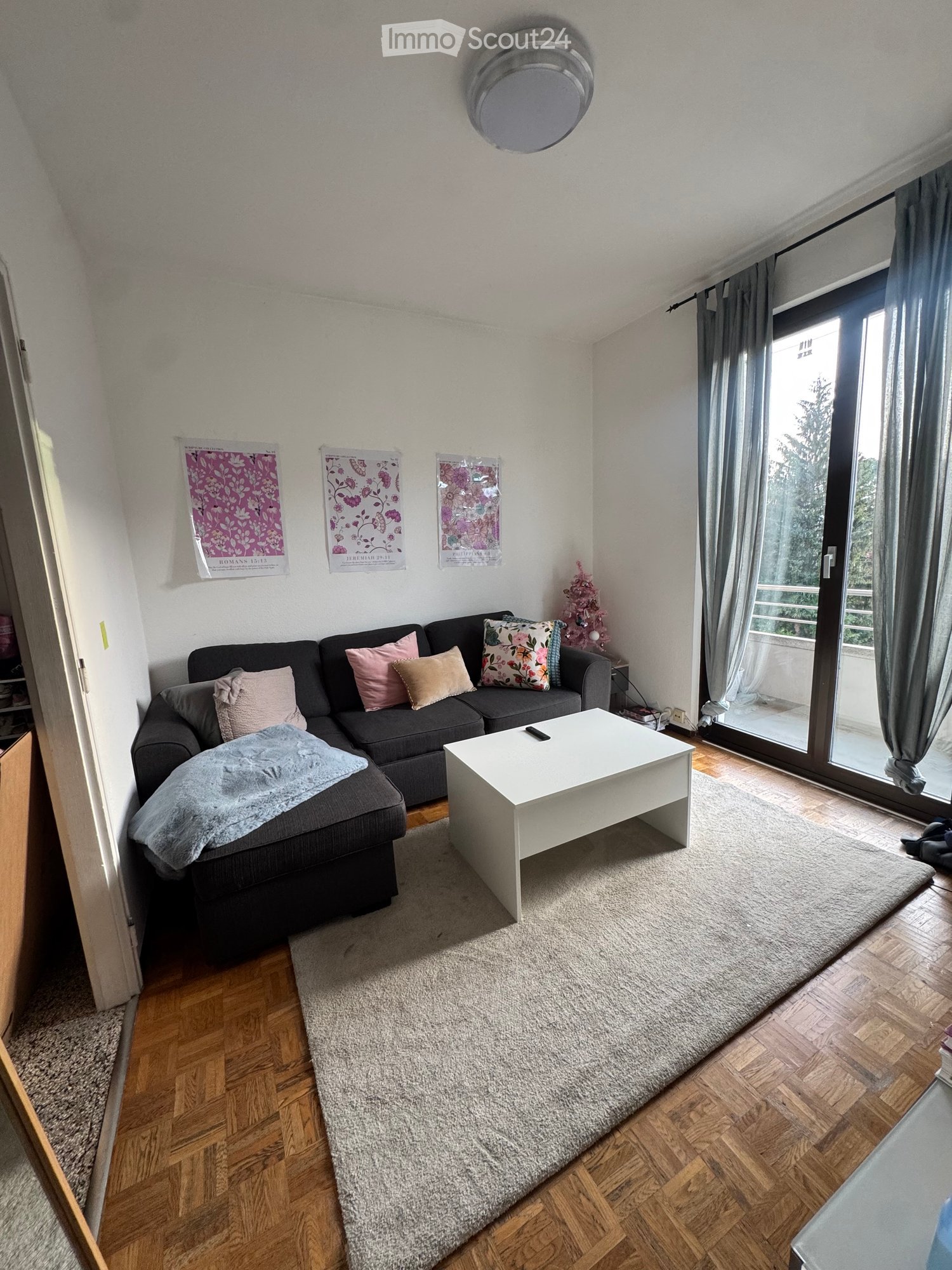 Modern living room with a grey sectional sofa, white coffee table, throw blanket, and decorative pillows. Features wooden flooring, large window with curtains, and posters on the wall.