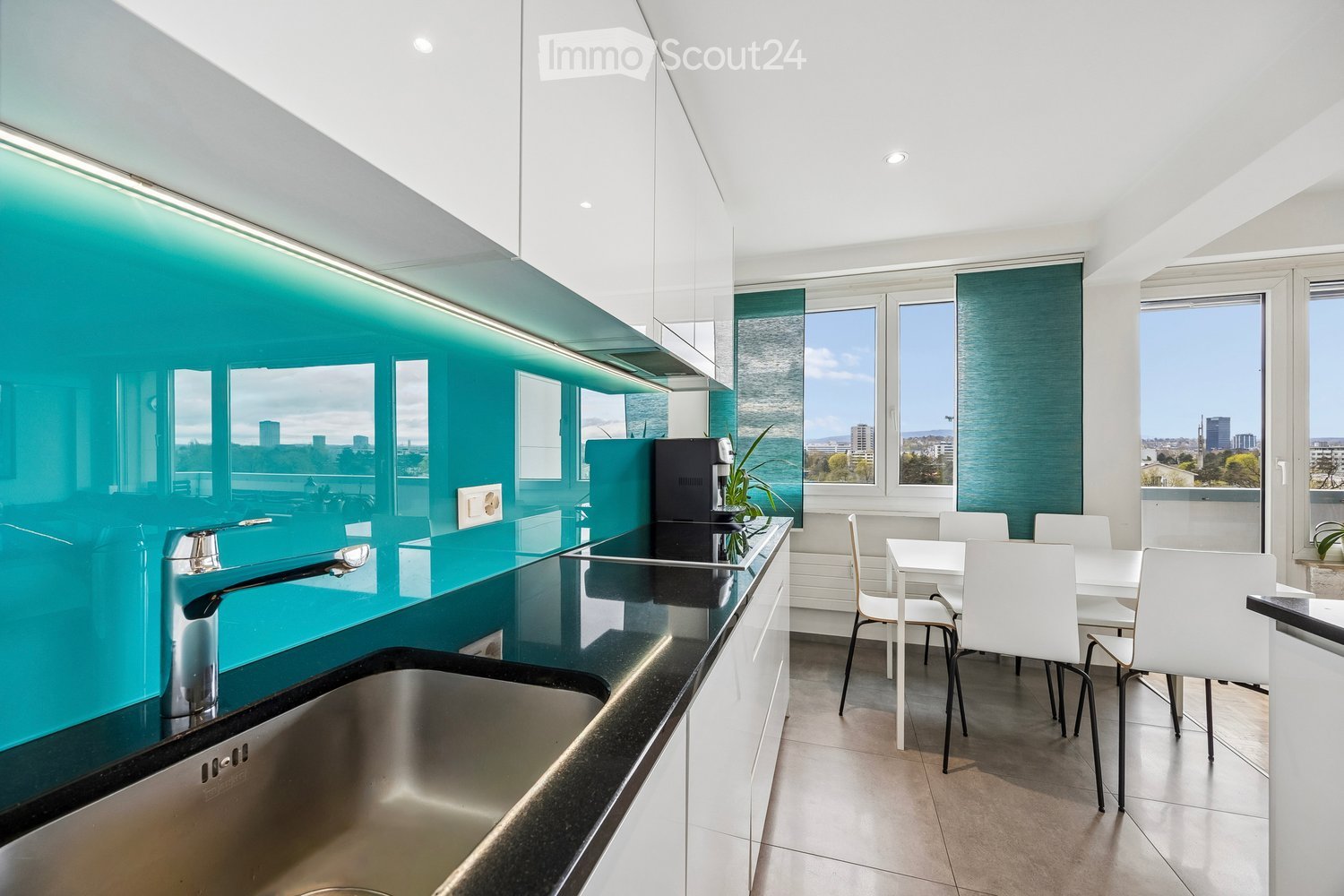 Modern kitchen with teal backsplash, stainless steel sink, white dining table, and chairs, overlooking a cityscape.
