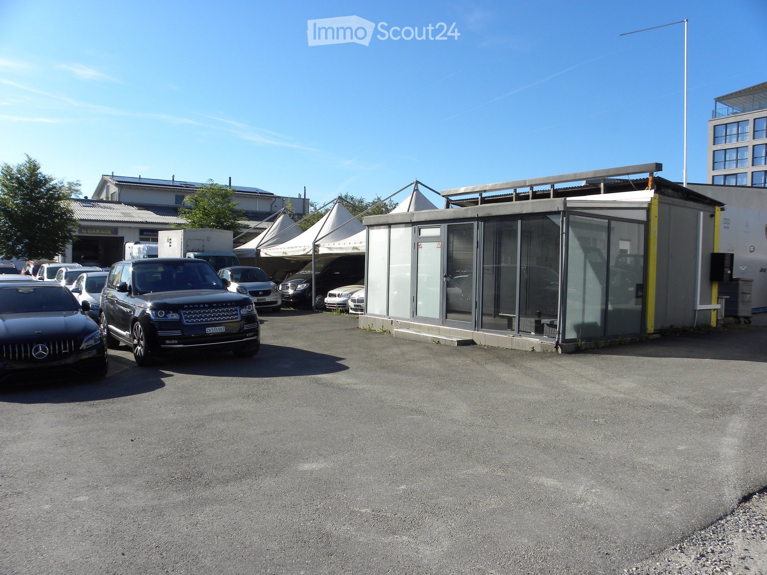 Outdoor parking area with several cars, a small garage, and a modern building with glass walls.