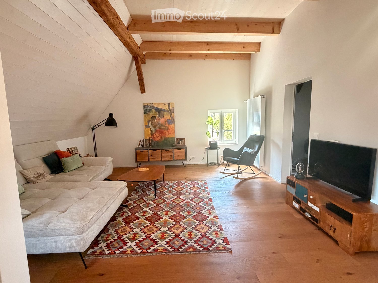 Bright living room with wooden floors, exposed wooden beams, a large sectional sofa, a coffee table, a rocking chair, a TV stand, a floor lamp, a side table with a plant, and a colorful rug.