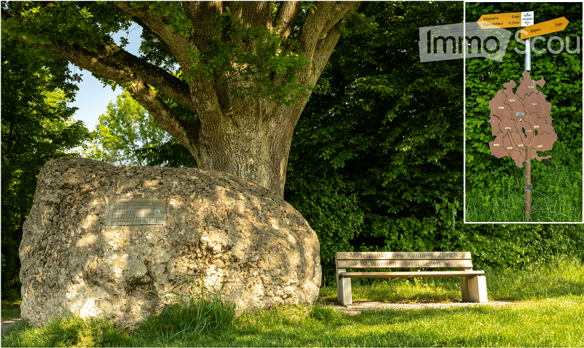 A large stone monument stands prominently in a grassy area, surrounded by lush greenery and trees. A wooden bench is positioned nearby, and a directional signpost is visible in the upper right corner, indicating nearby locations.
