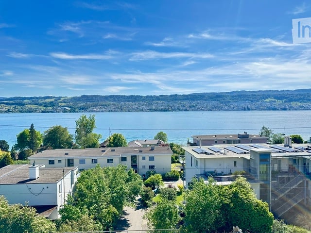 Aerial view of a residential complex with multiple buildings, surrounded by trees, overlooking a large body of water and distant hills.