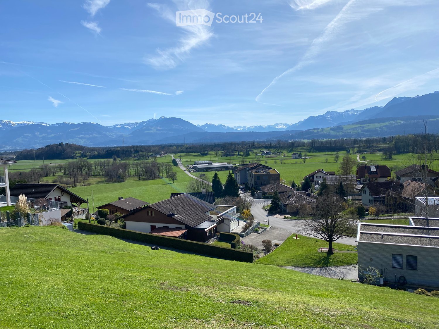 A panoramic view of a rural village with houses, green fields, and distant mountains under a clear blue sky.