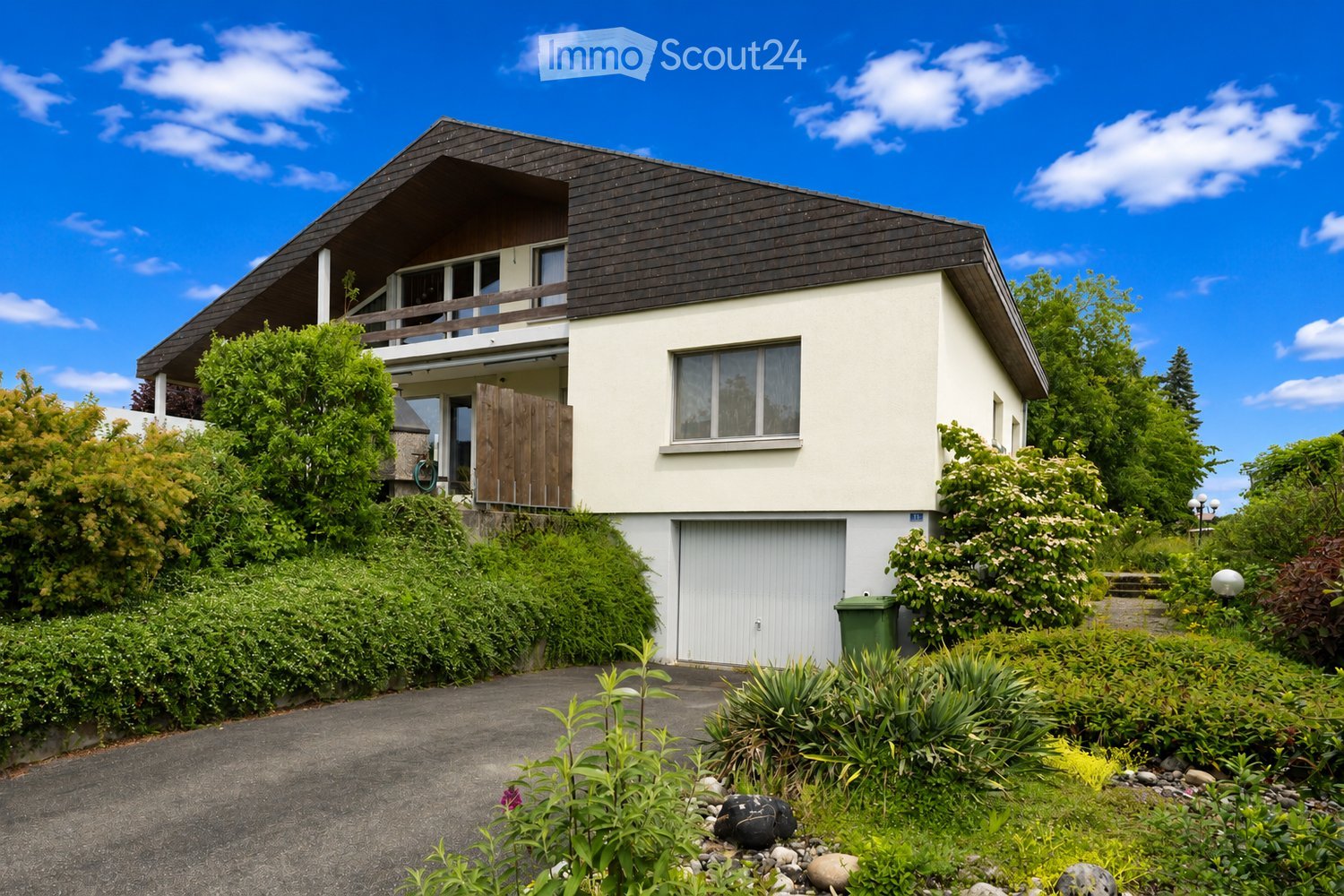 Two-story house with a dark roof, white walls, and a balcony on the upper level. The house features a garage, surrounded by lush greenery and a well-maintained garden.