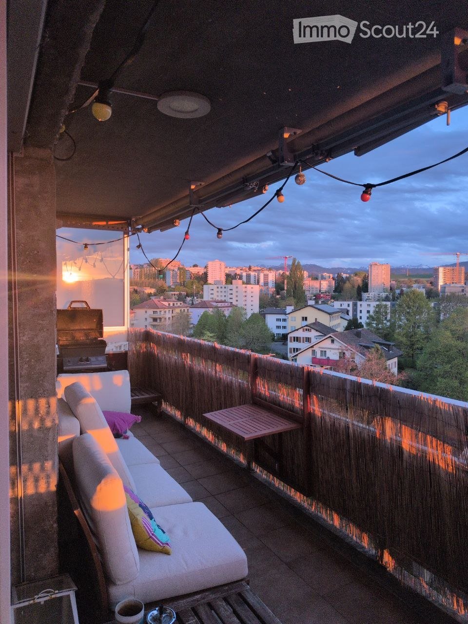 Balcony with a white sofa, small table, grill, and string lights, overlooking a cityscape at sunset.