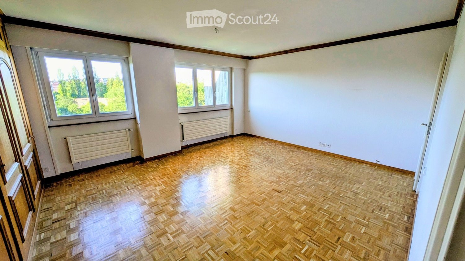 Empty room with wooden parquet flooring, white walls, and large windows offering a view of greenery outside. Radiators are visible beneath the windows.