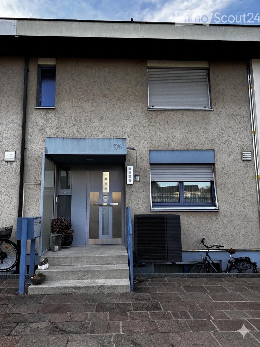Modern building entrance with a grey facade, large windows, and a small staircase leading to the door. There are bicycles parked nearby and a small potted plant on the steps.