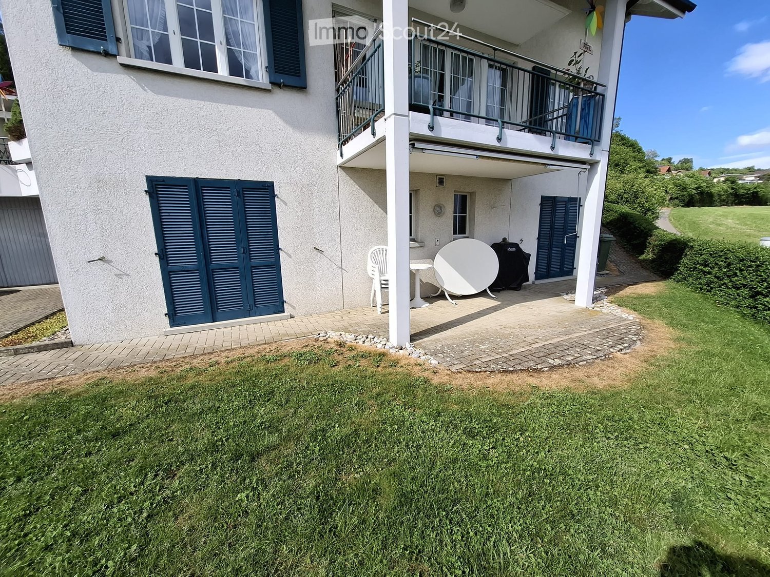A well-maintained garden with a paved patio area, featuring a white table, a chair, and a satellite dish. The house has multiple windows with shutters and a balcony with railings.