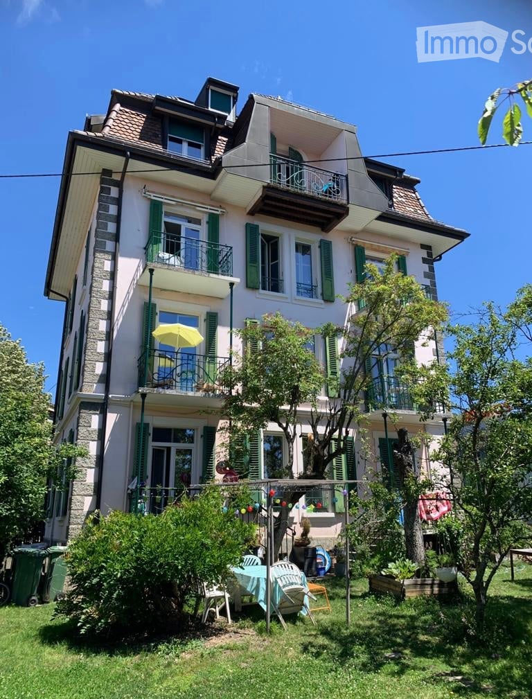 A multi-story house with green shutters, balconies, and a well-maintained garden with outdoor furniture.