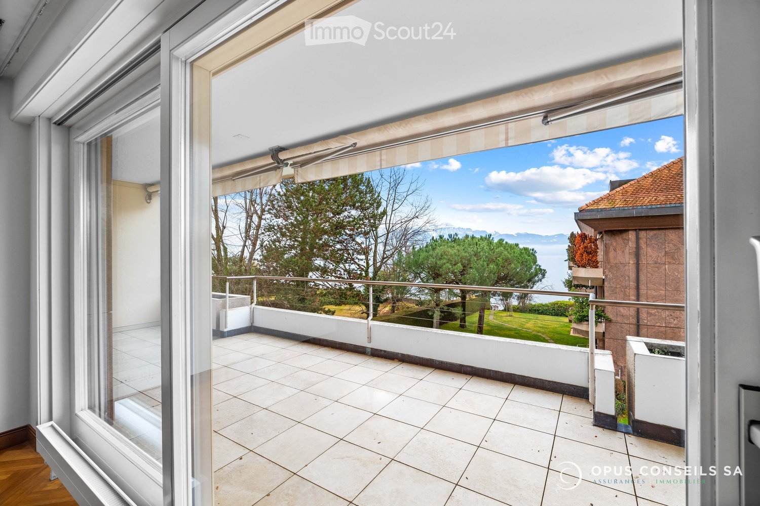 Spacious balcony with tiled flooring, white railings, and a scenic view of a green lawn, trees, and a body of water under a clear blue sky.