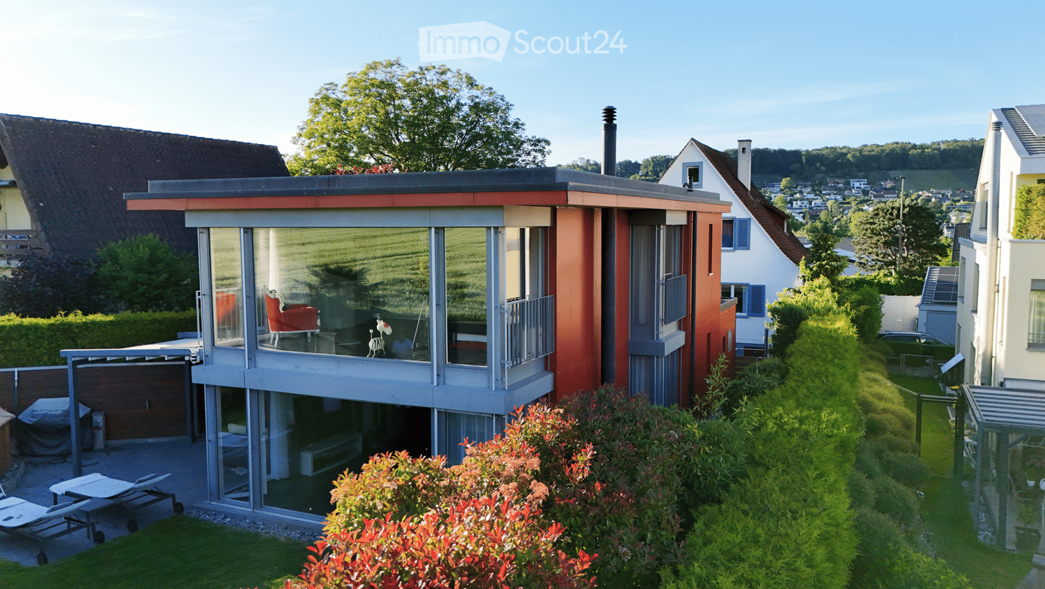 Modern two-story house with large glass windows and a red and white facade, surrounded by greenery and neighboring houses.