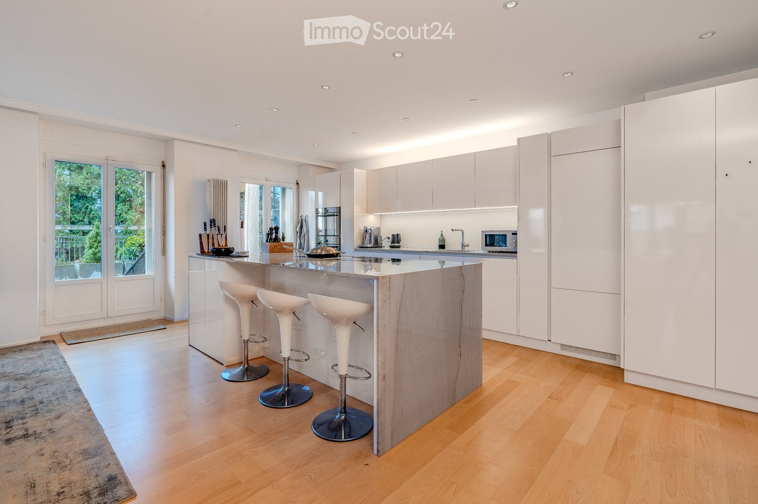Modern kitchen with white cabinets, a central island with bar stools, and various appliances including a microwave and blender. The space features wooden flooring and large windows with a view of greenery.