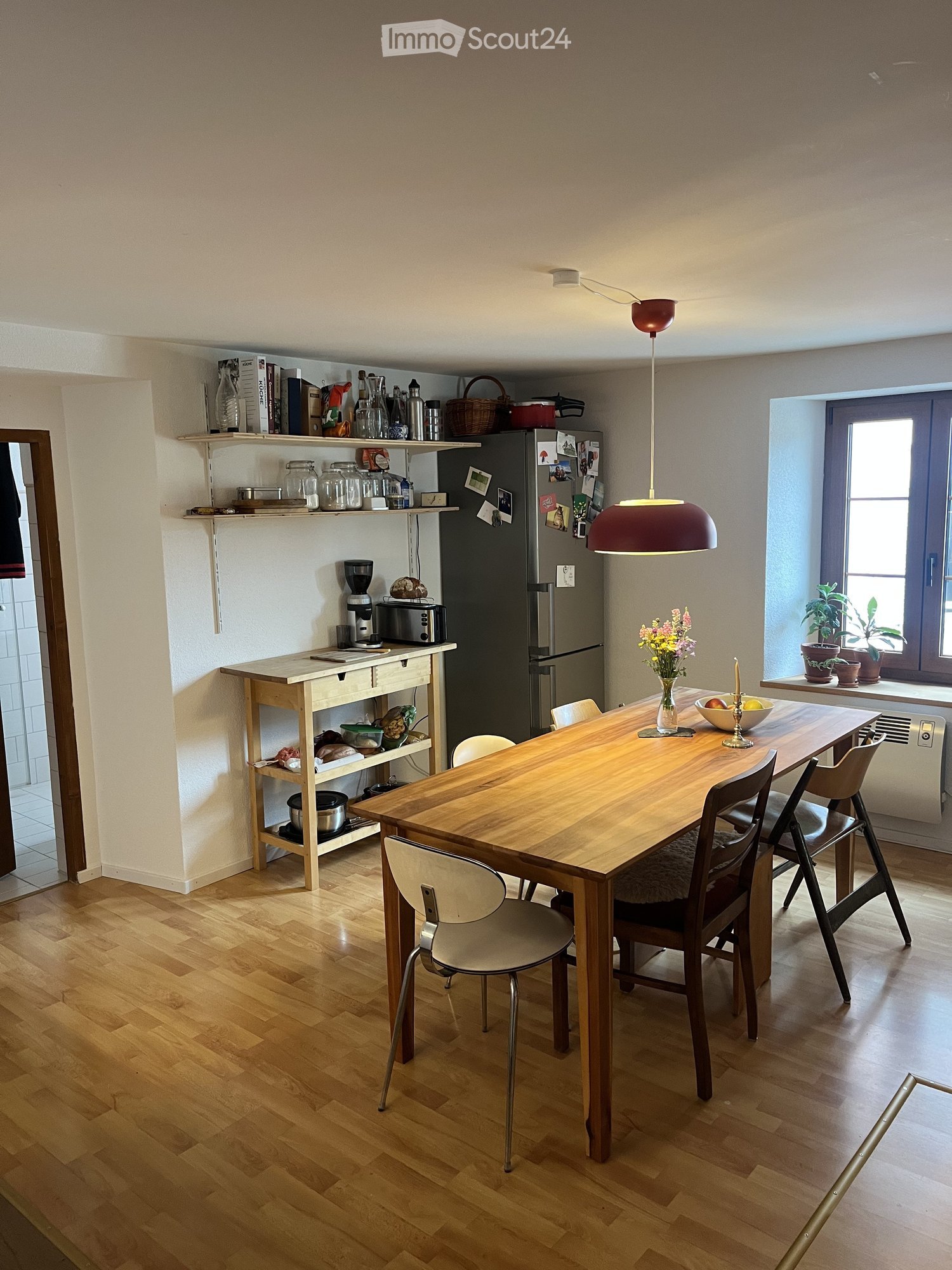 Bright kitchen and dining area with wooden flooring, a long wooden dining table surrounded by chairs, a refrigerator, coffee maker, toaster, shelves with jars and bottles, and a vase with flowers on the table.