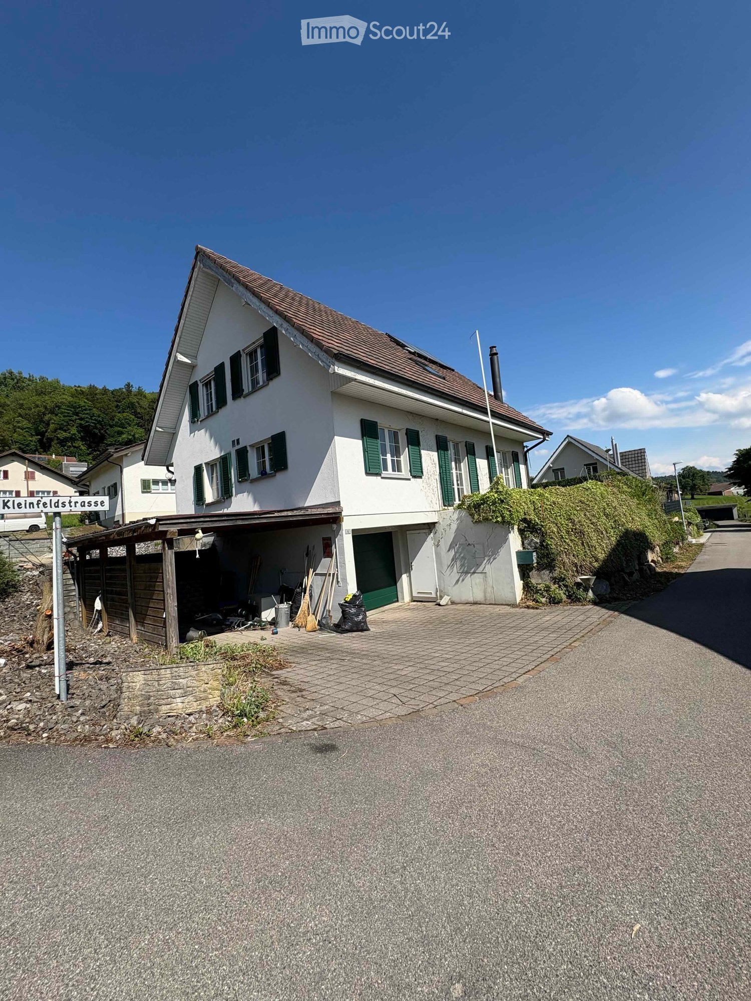 Two-story white house with green shutters, brown roof, and attached garage. The house is situated on a raised ground floor with a paved driveway and a small garden area.