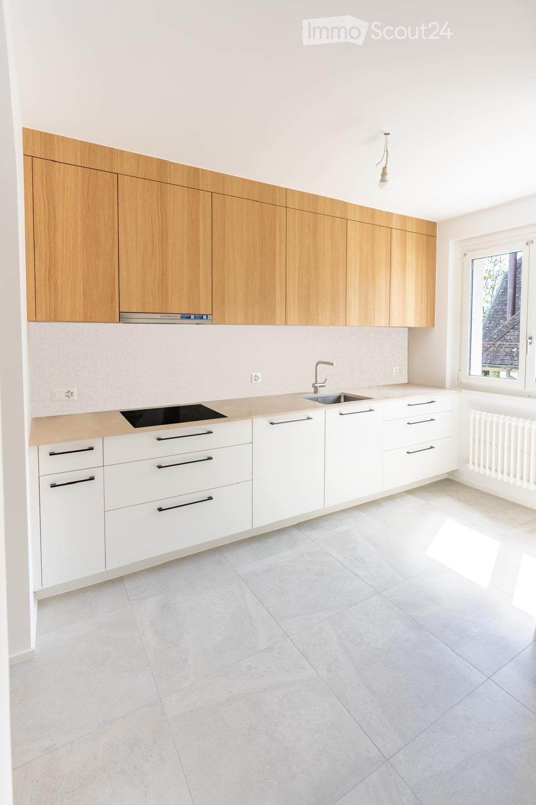 Modern kitchen with light wooden upper cabinets, white lower cabinets, a sink, and a stove. The space features a light-colored tiled floor and ample natural light from a large window.