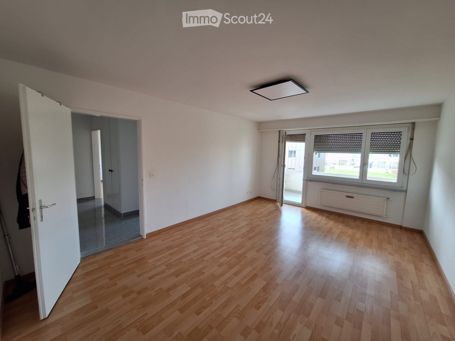Empty room with wooden flooring, white walls, and large windows. There is a door leading to another room and a radiator under the windows.