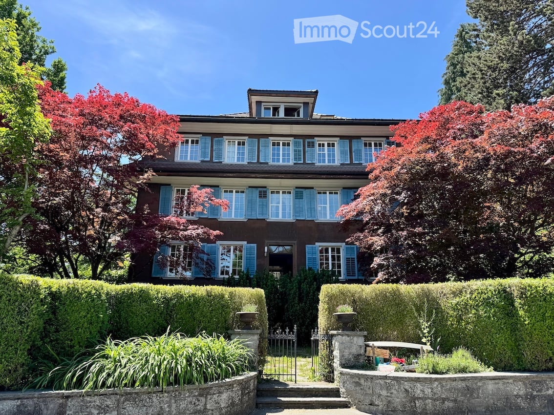 A two-story house with a dark brown exterior and light blue shutters, surrounded by well-maintained hedges and trees.