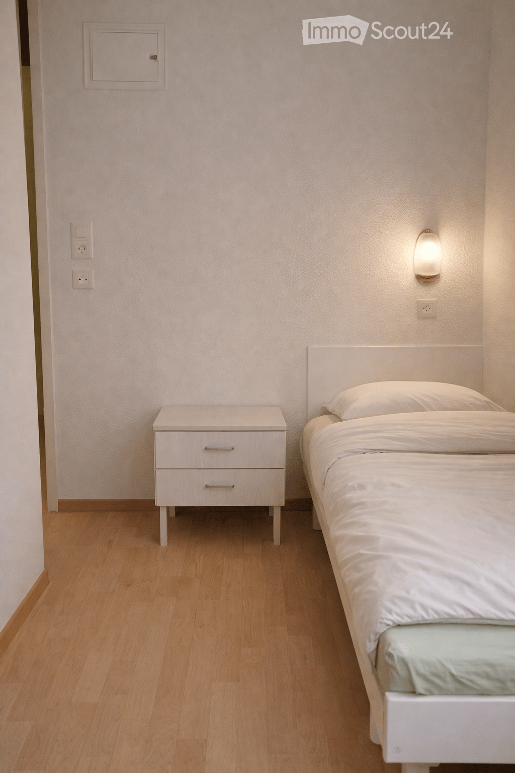 Bedroom with wooden floor, white walls, a bed with white sheets and a white pillow, a small white nightstand with drawers, a wall lamp, and electrical outlets.