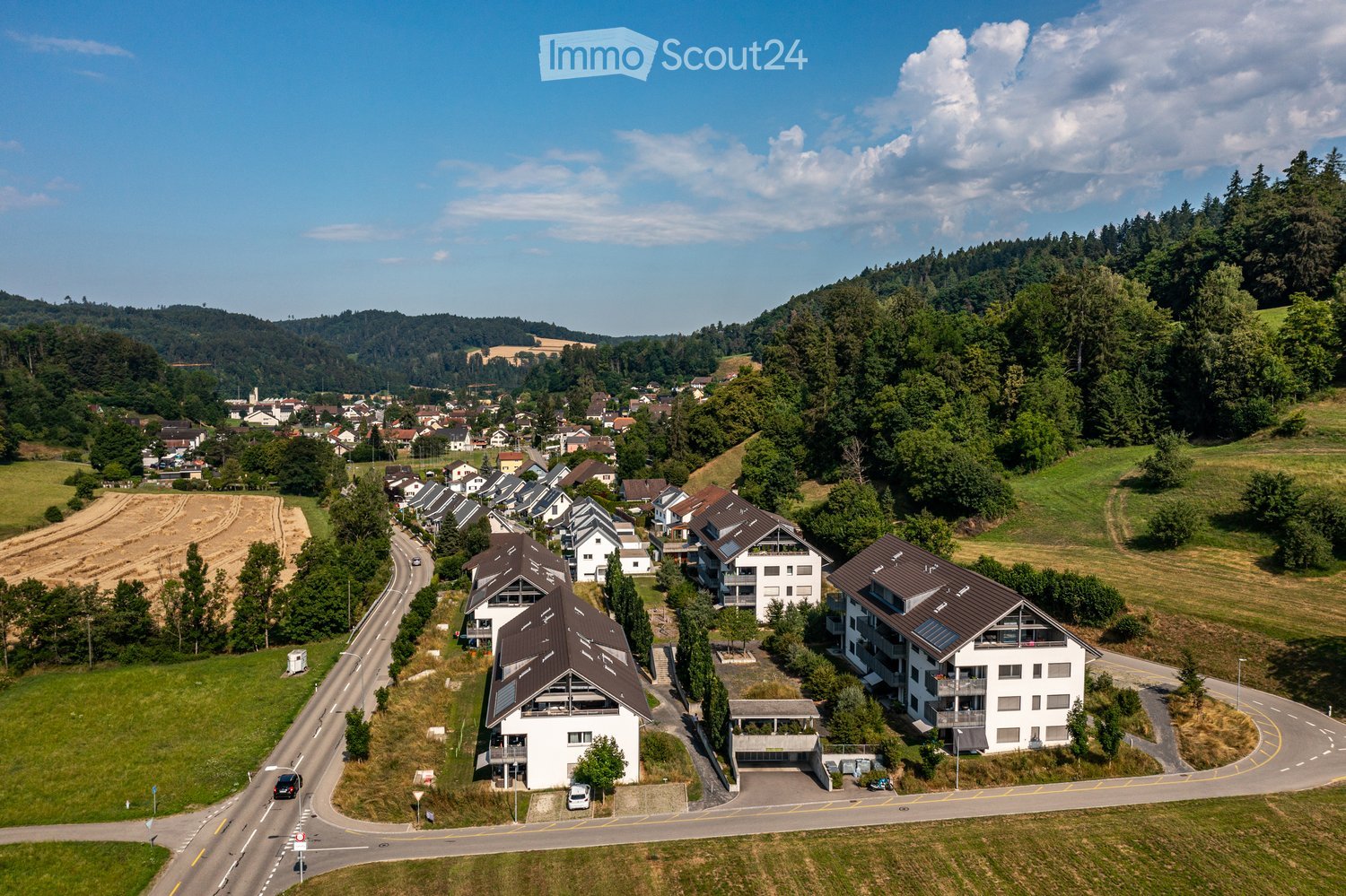 Aerial view of a residential area with multiple houses, surrounded by greenery and hills in the background.
