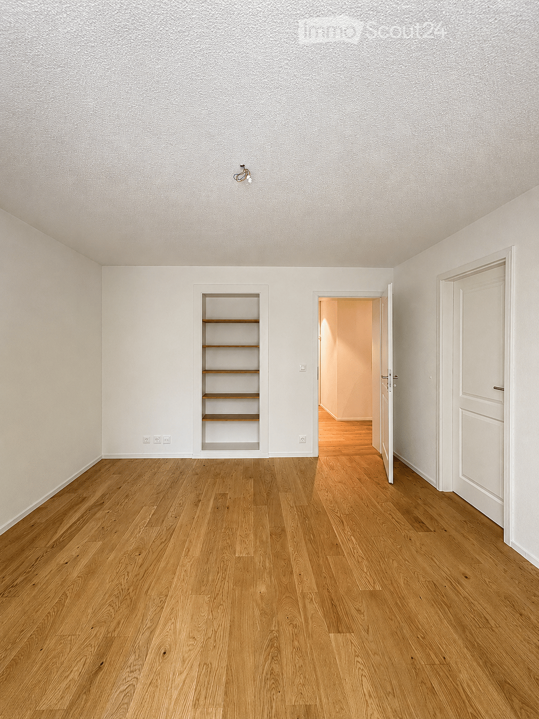 Empty room with light wooden flooring, white walls, and a built-in wooden shelf unit. The room features a door leading to another space and a visible electrical outlet on the wall.
