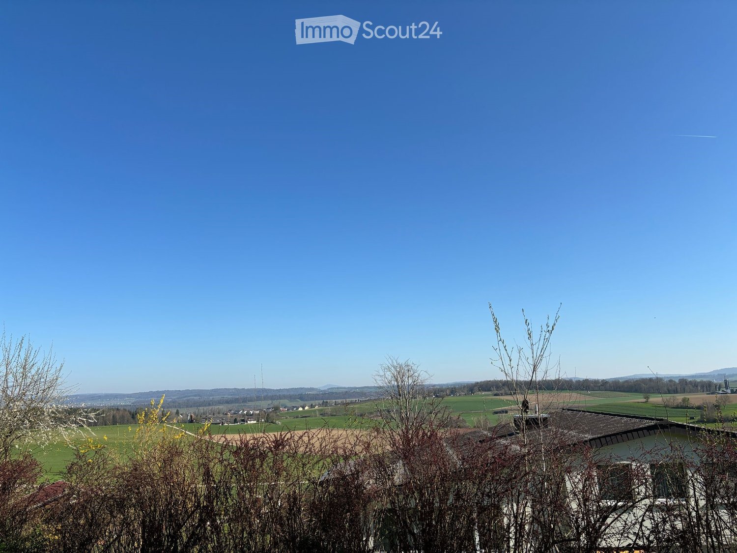A panoramic view of a rural landscape with fields, scattered houses, and distant hills under a clear blue sky.