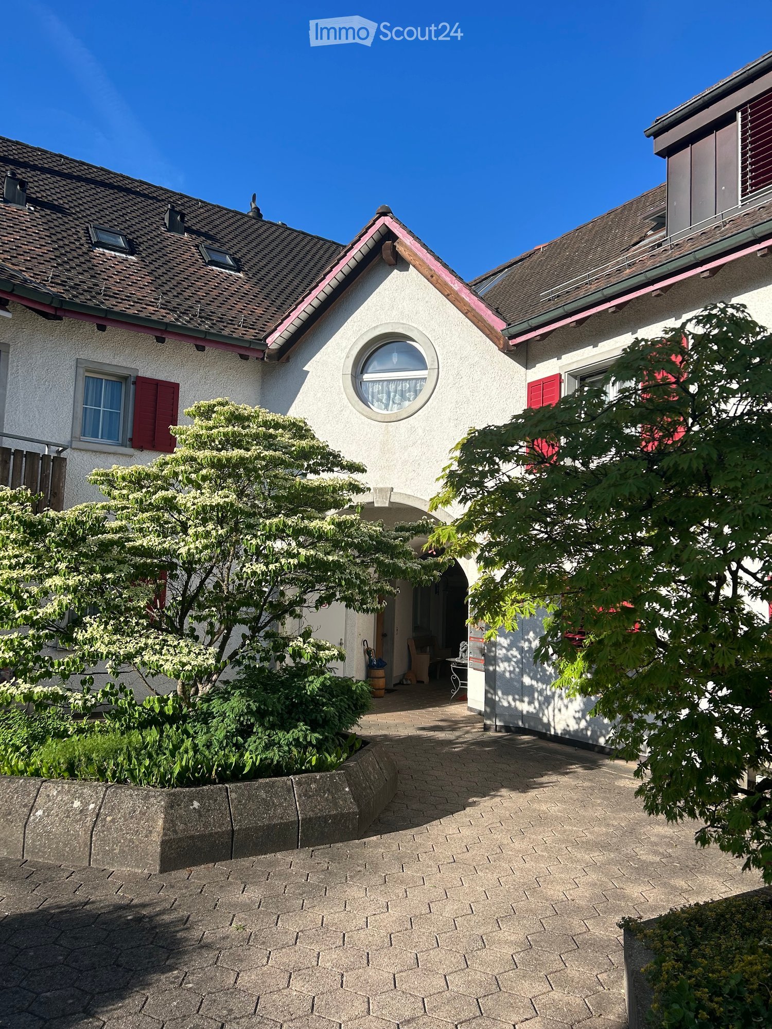 Two-story house with white walls, red shutters, and a tiled roof. Features include a circular window, a small porch with a table and chairs, and a well-maintained garden with trees and shrubs.