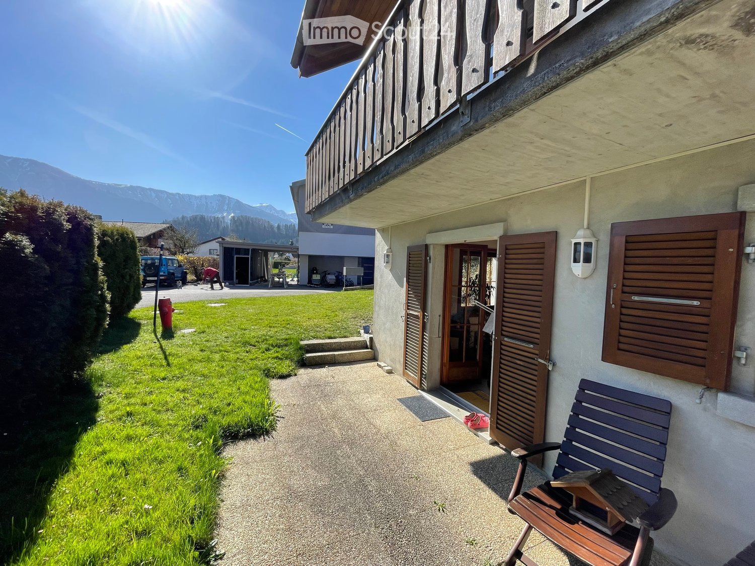 Garden area with a wooden bench, grassy lawn, and a pathway leading to the house entrance. The house features a balcony and wooden shutters.