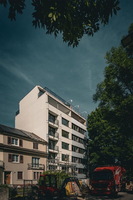 Modern multi-story apartment building with balconies, surrounded by trees and adjacent to a smaller house. Construction vehicles are visible in the foreground.