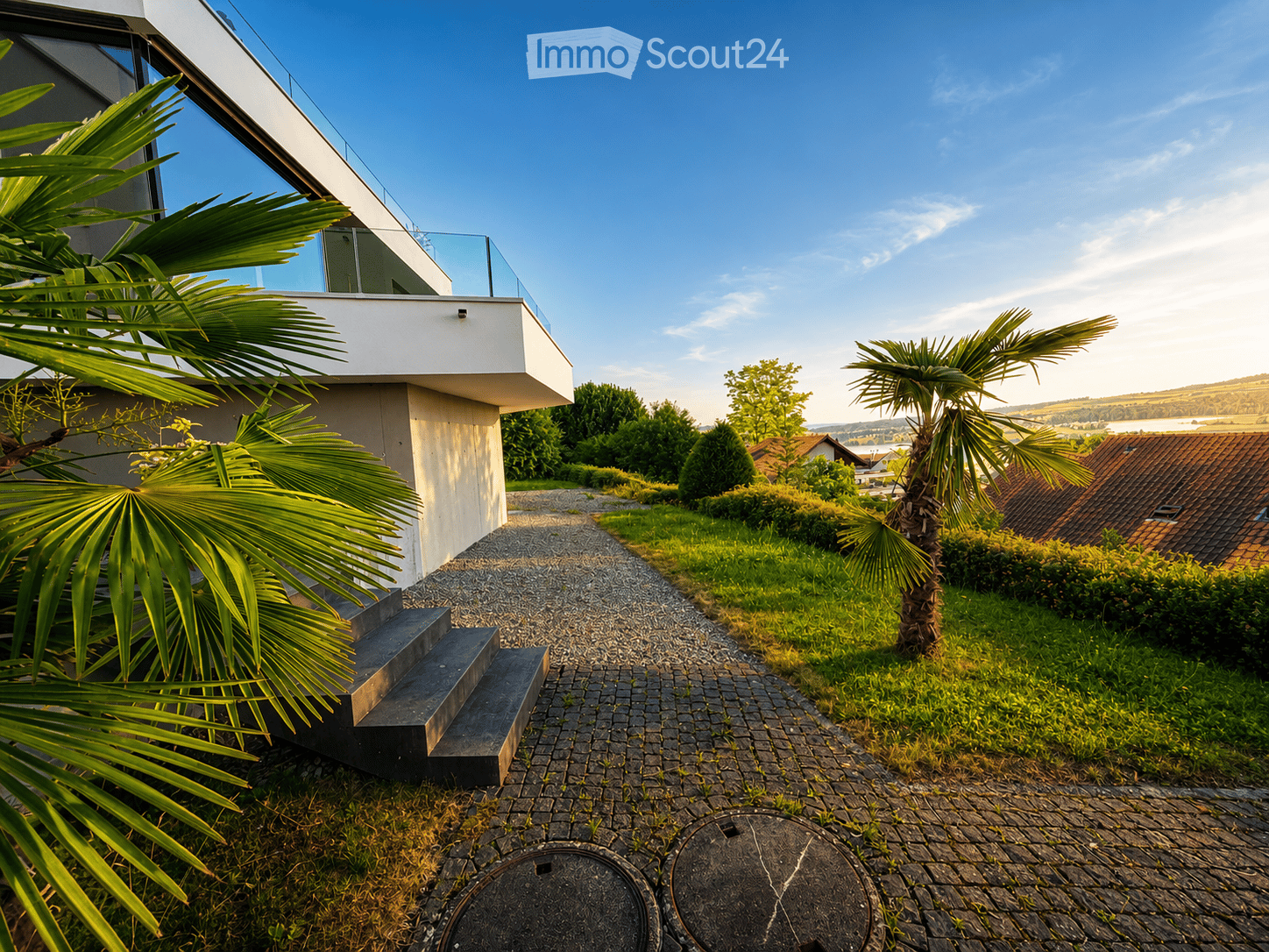 Modern house exterior with a raised ground floor, surrounded by a well-maintained garden with palm trees, gravel pathways, and a scenic view of the countryside.