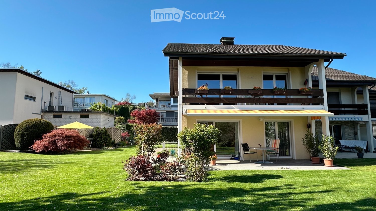 Two-story house with a balcony, surrounded by a well-maintained garden with lawn, shrubs, and potted plants. The house features large windows and a covered patio area with outdoor furniture.