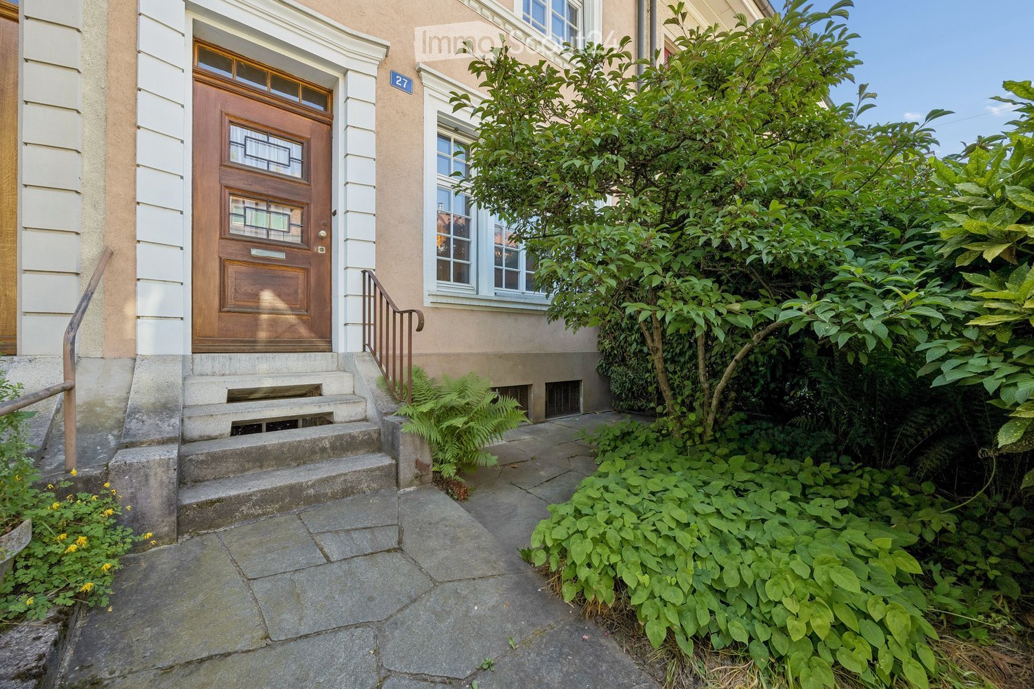 Front entrance of a two-story house with a wooden door, steps, and lush greenery surrounding the pathway.