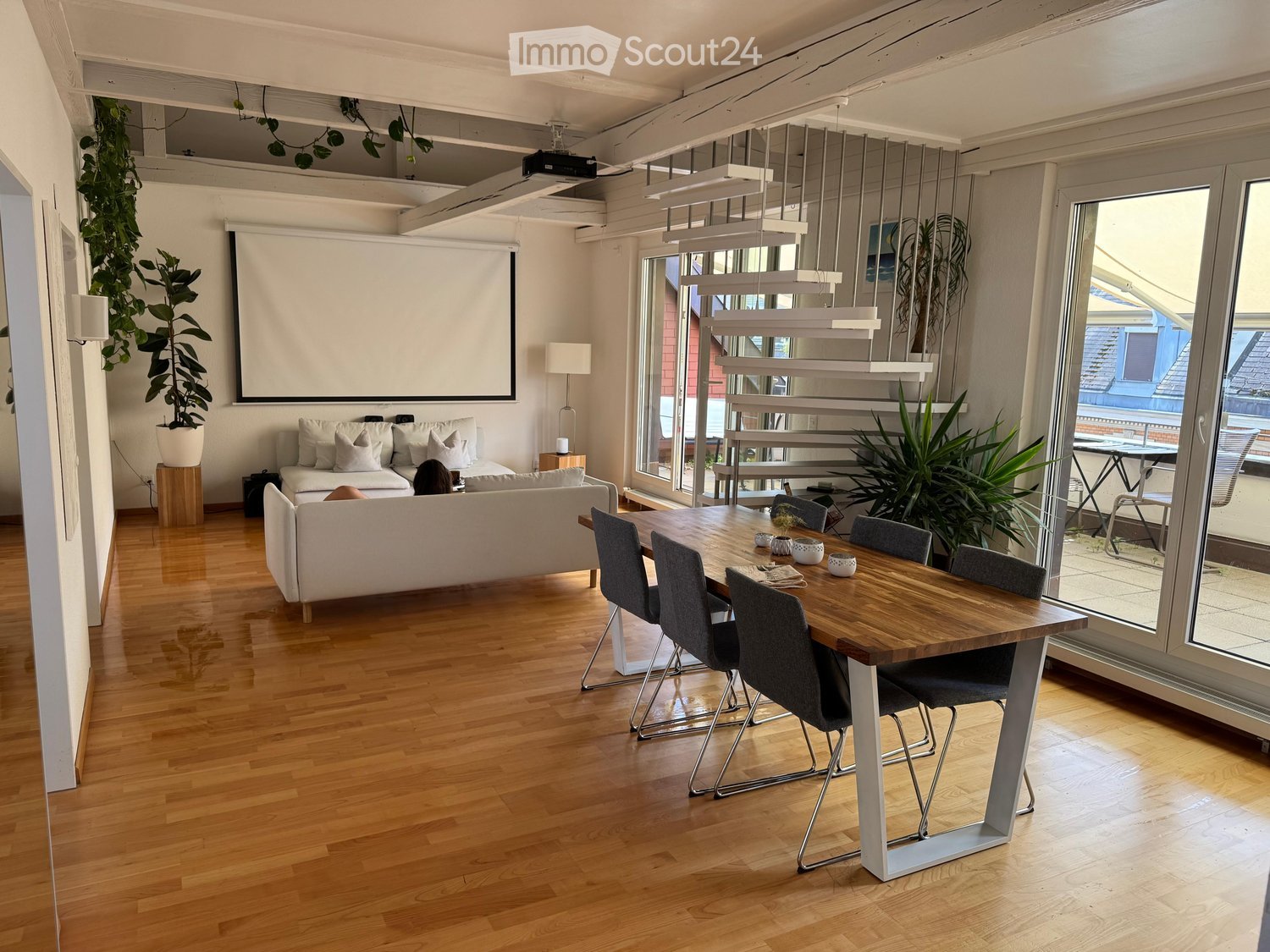 Open-plan living and dining area with wooden flooring, white sofa, dining table with chairs, pendant lights, and large windows leading to a balcony.