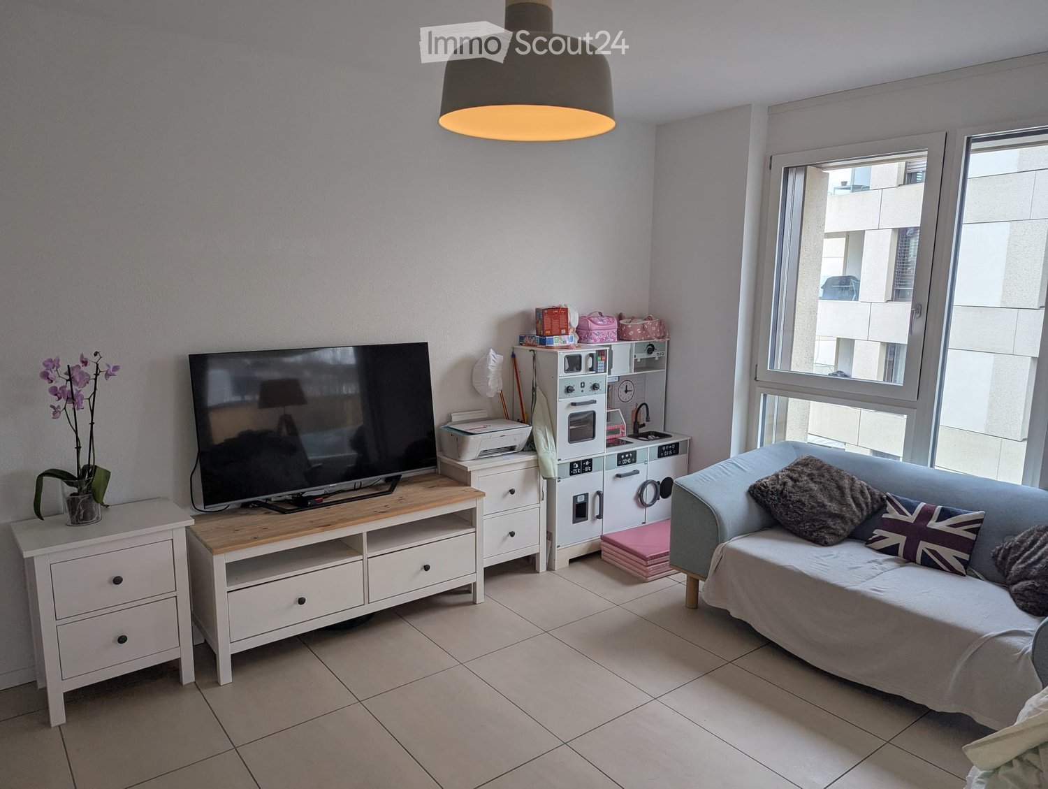 A modern living room with a sofa, television, wooden coffee table, and a mini fridge. The room features large windows with a view of the building outside, a hanging pendant light, and a small side table with a potted plant.