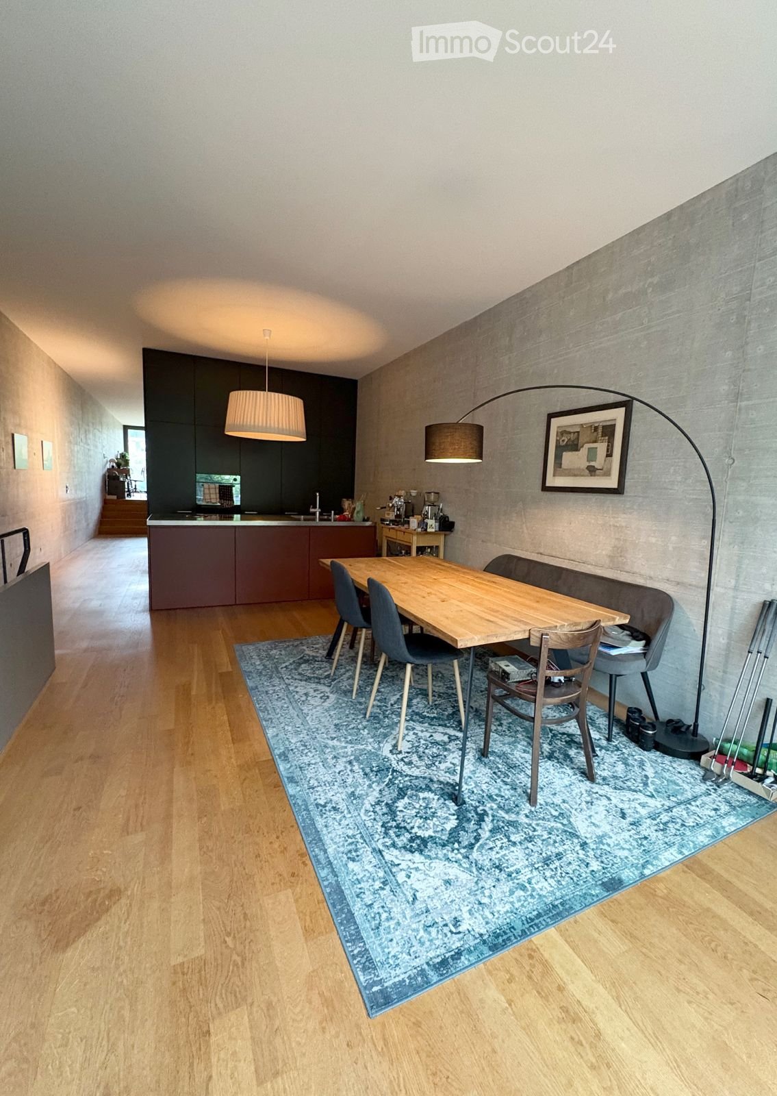 Dining area with a wooden table, four chairs, a sofa, a pendant light, a floor lamp, a framed picture, and a kitchen area with cabinets and a coffee machine.