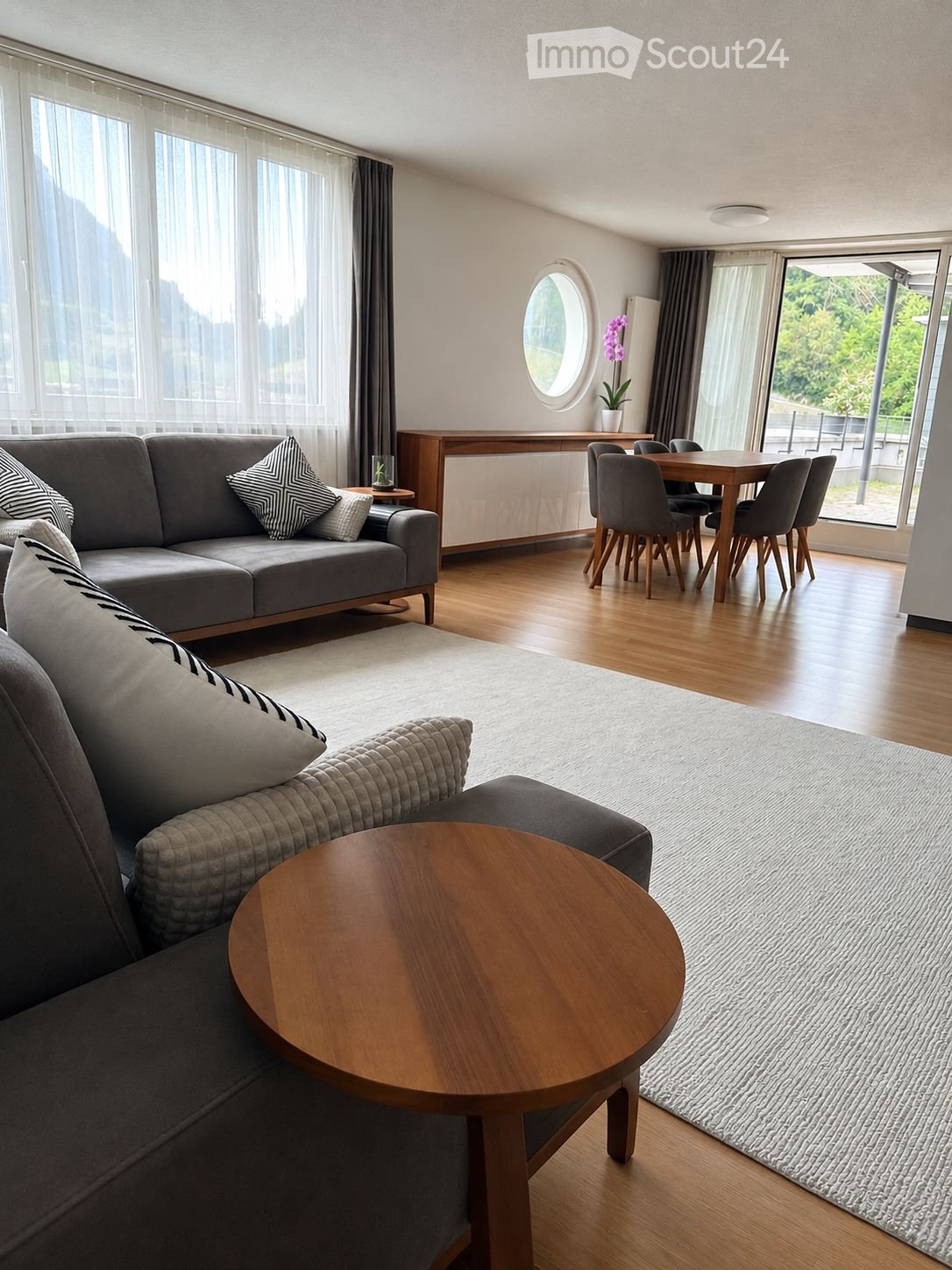 Modern living room with grey sofa, wooden coffee table, dining area with table and chairs, and large windows with a view.