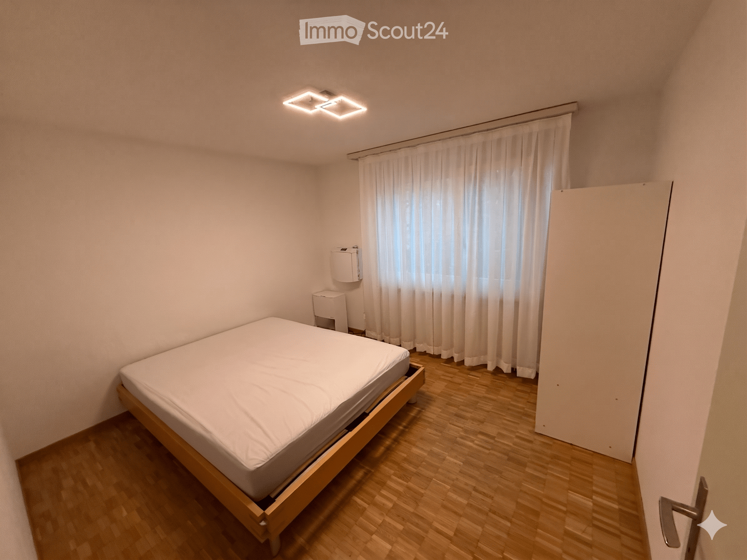 Minimalist bedroom with a wooden bed frame, white bedding, a window with white curtains, and a radiator on the wall.