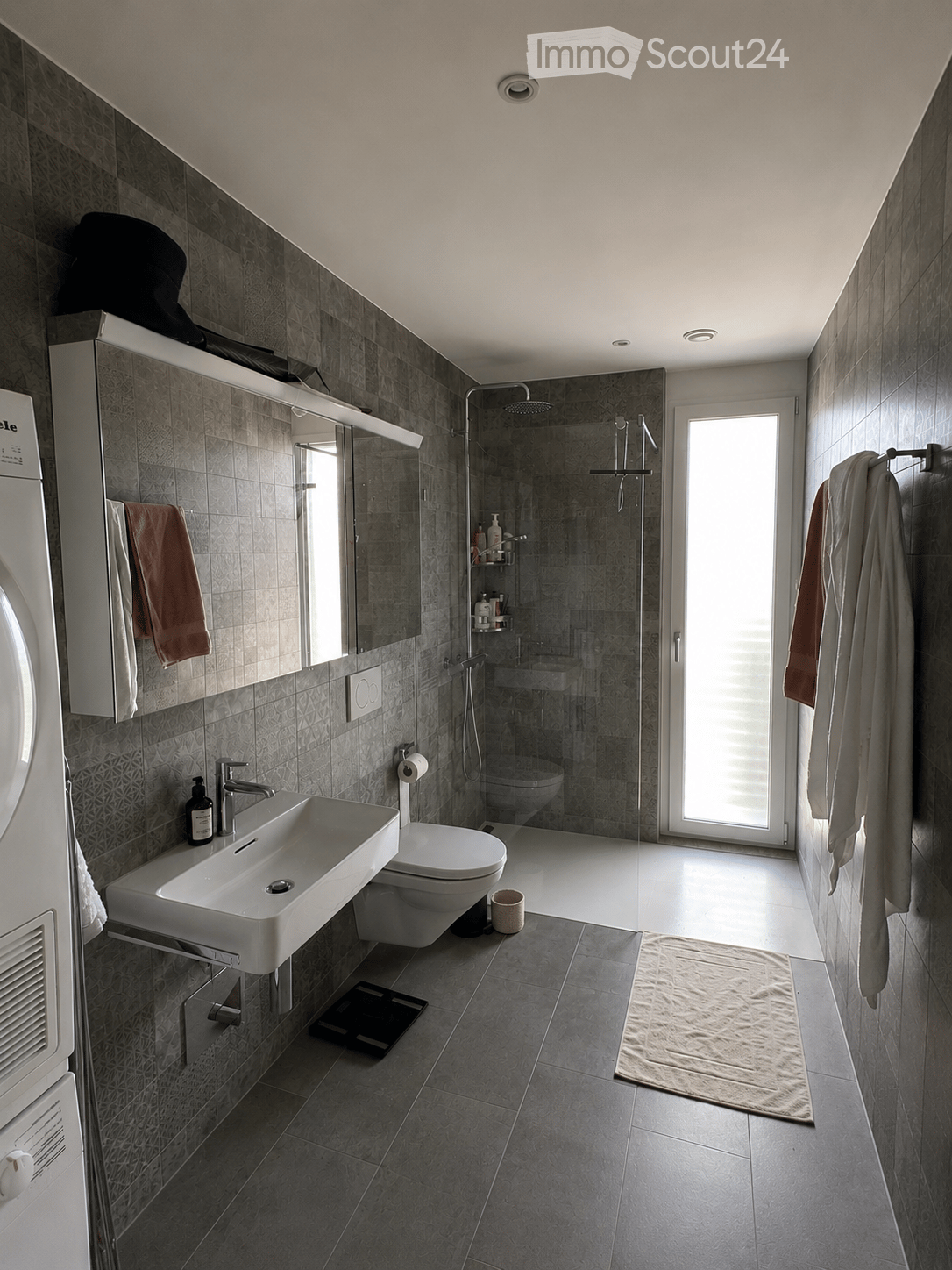 Modern bathroom with grey tiles, a white sink, toilet, shower, and a large mirror. Towels are hanging on the wall, and a small rug is placed on the floor.