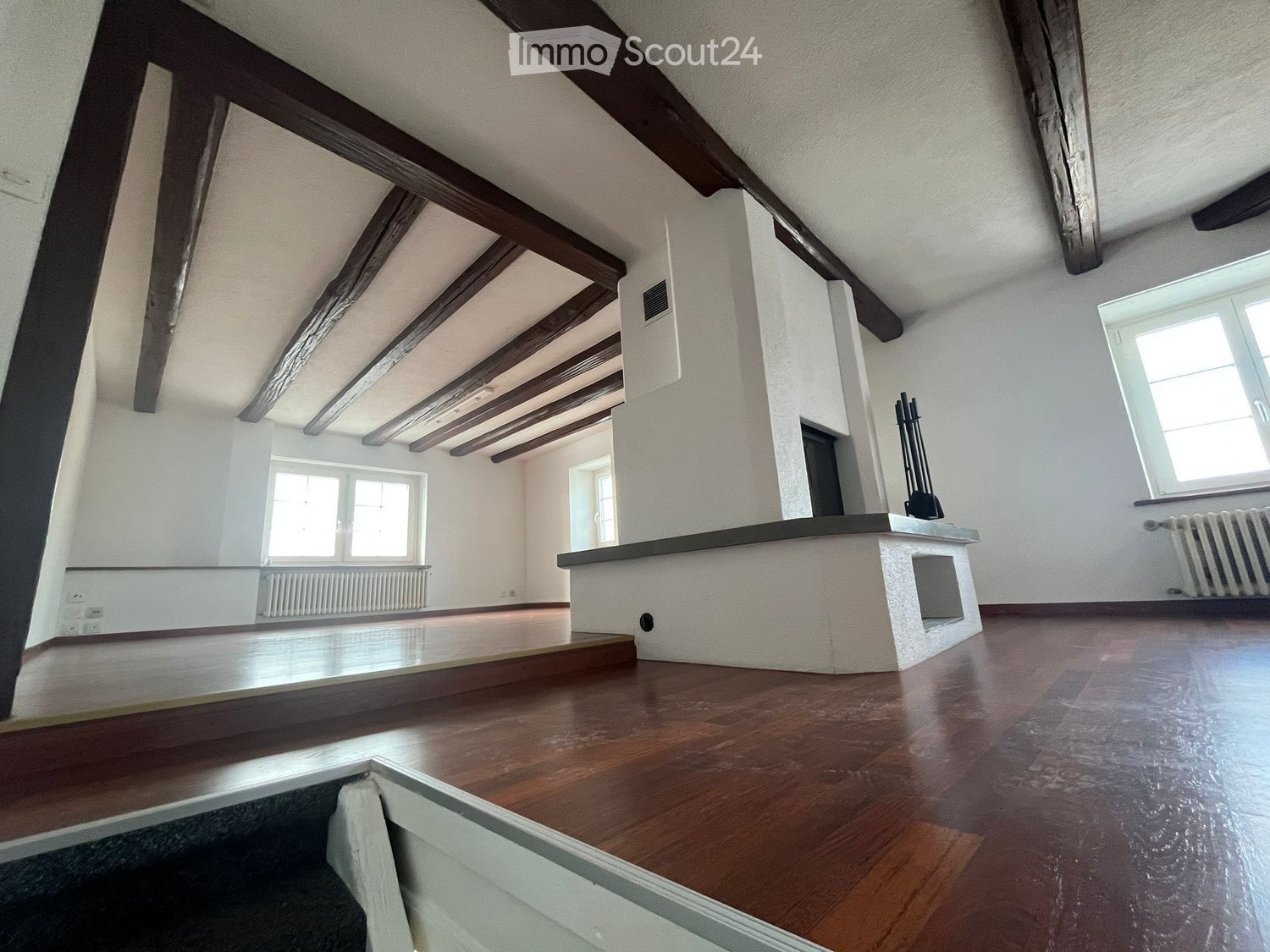 An empty room with wooden floors, exposed wooden beams on the ceiling, white walls, and large windows allowing natural light. There is a fireplace with a black mantelpiece and radiators along the walls.