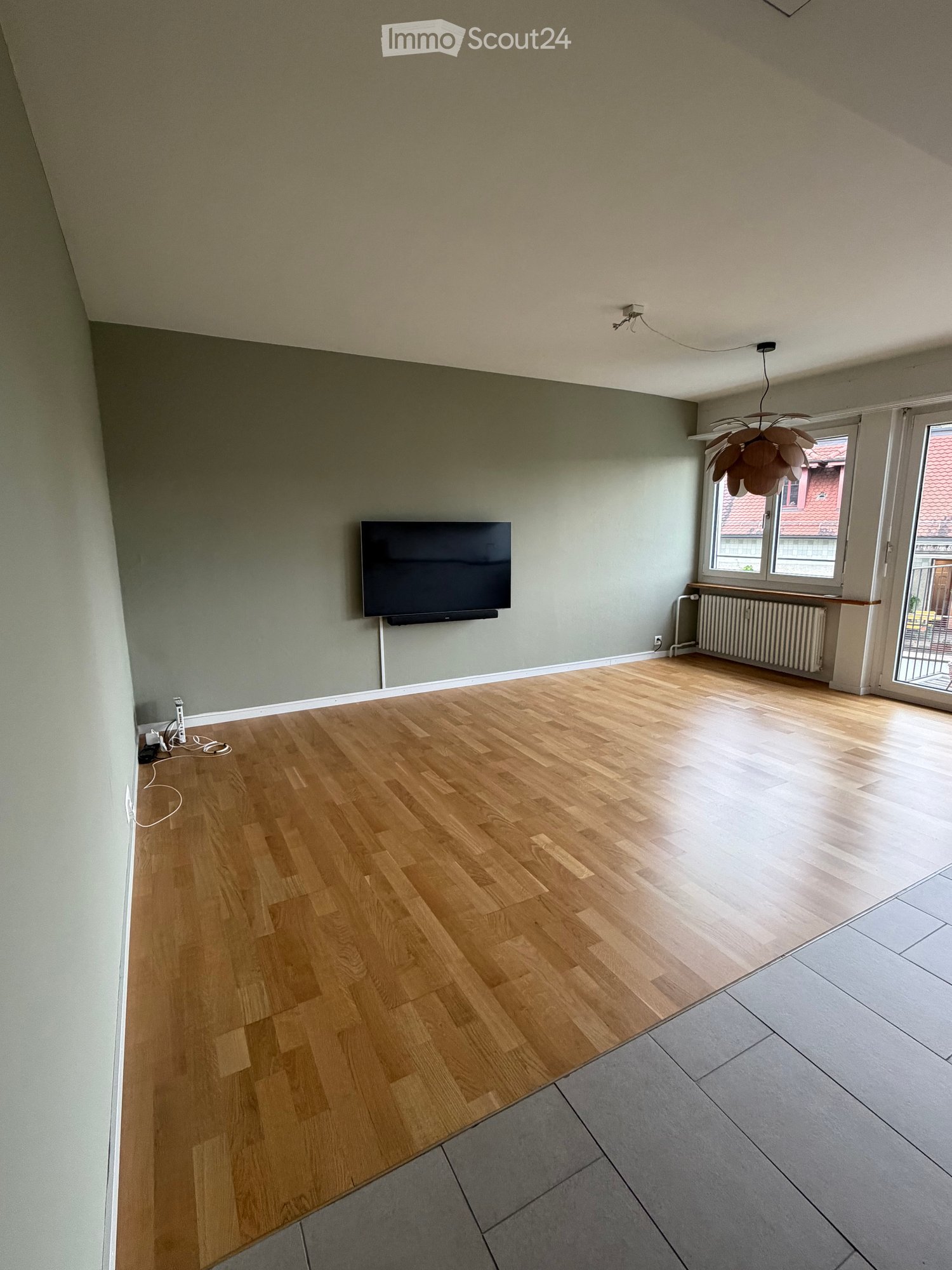 Empty room with wooden flooring, light green walls, and a mounted TV on one wall. There is a radiator under a window and a hanging light fixture above.