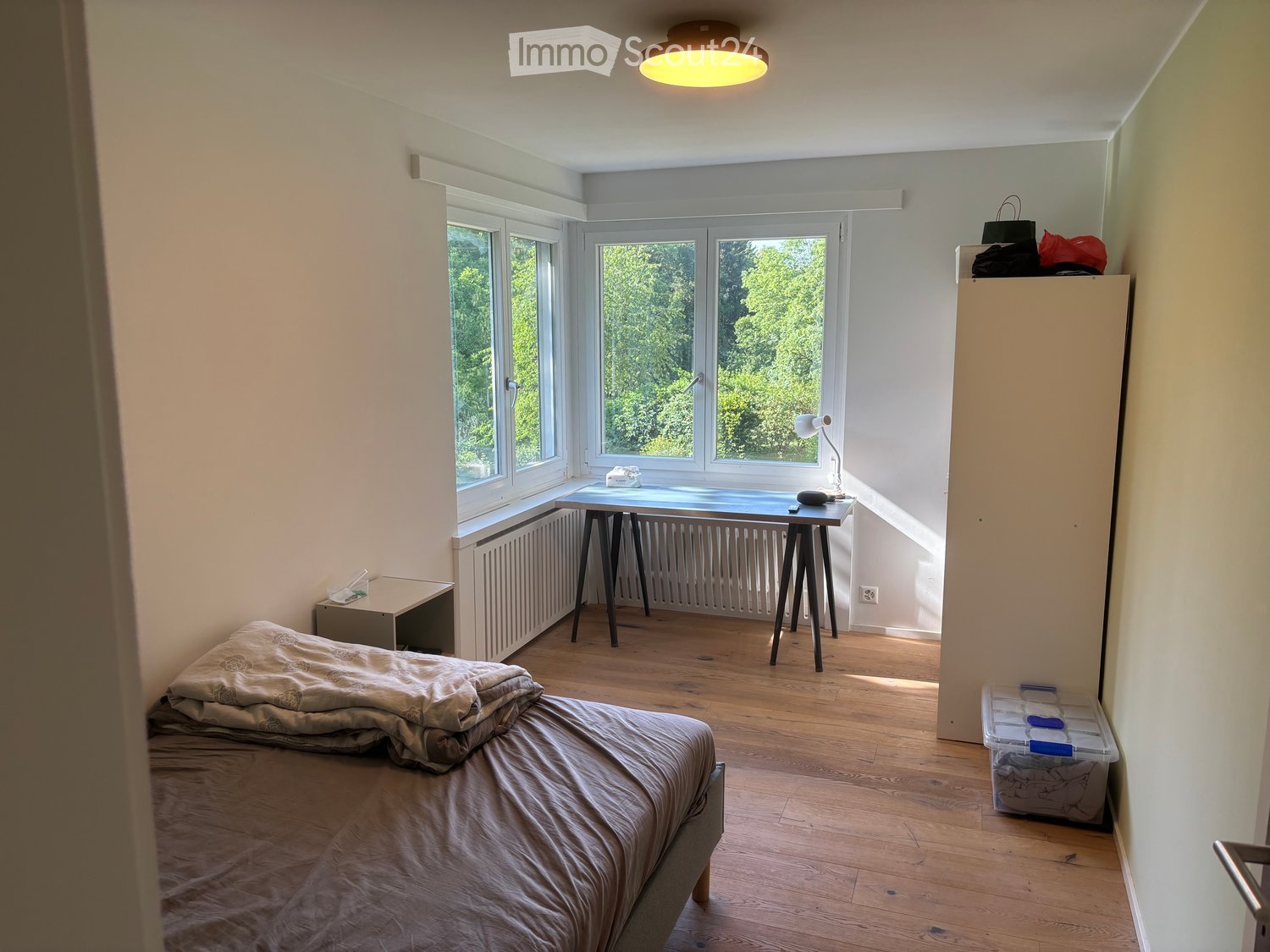 Cozy bedroom with wooden flooring, large windows, a bed with bedding, a small table with a lamp, and storage items.