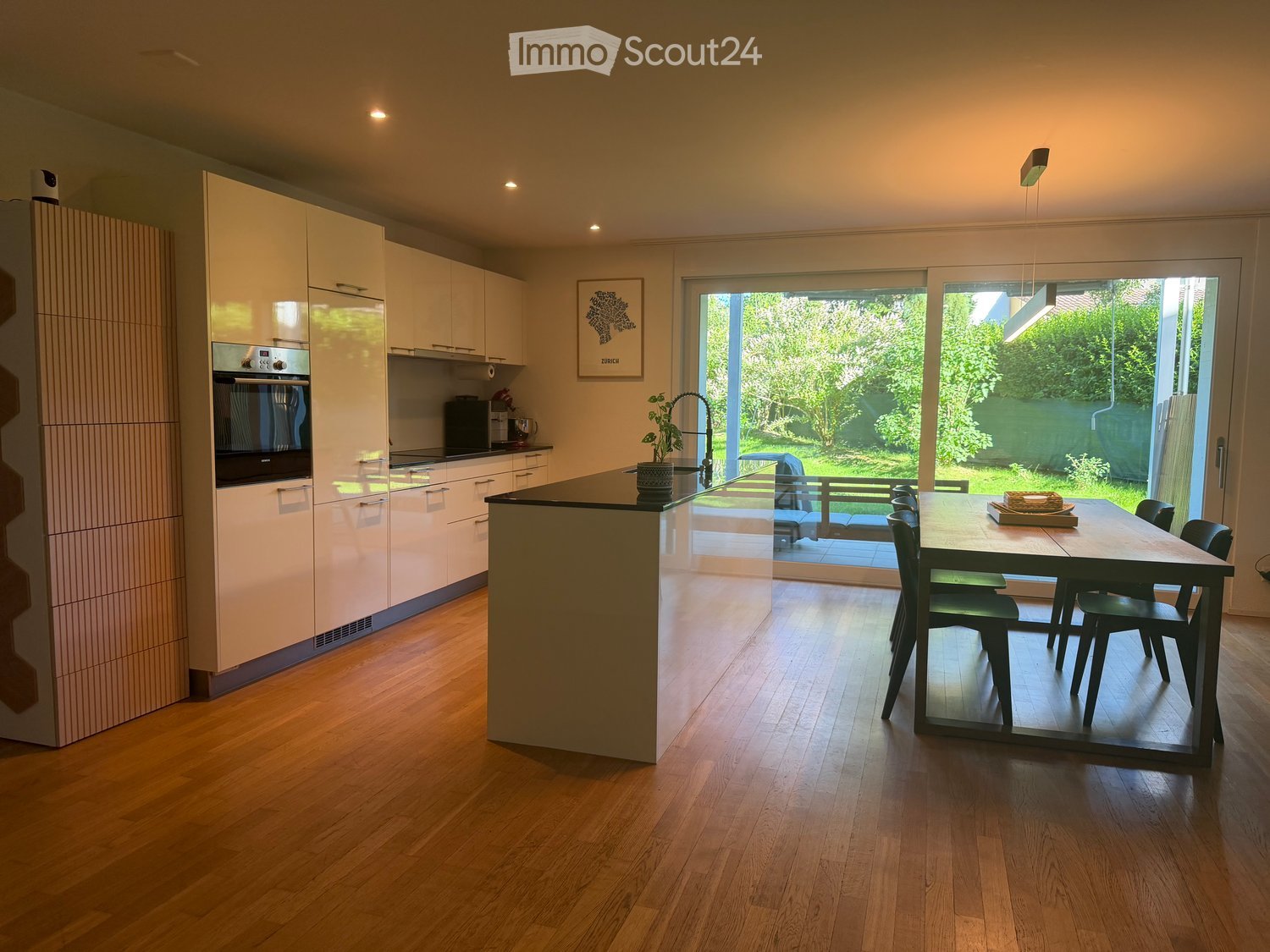 Modern kitchen with light-colored cabinetry, an island, a dining table with chairs, and a potted plant on the island.