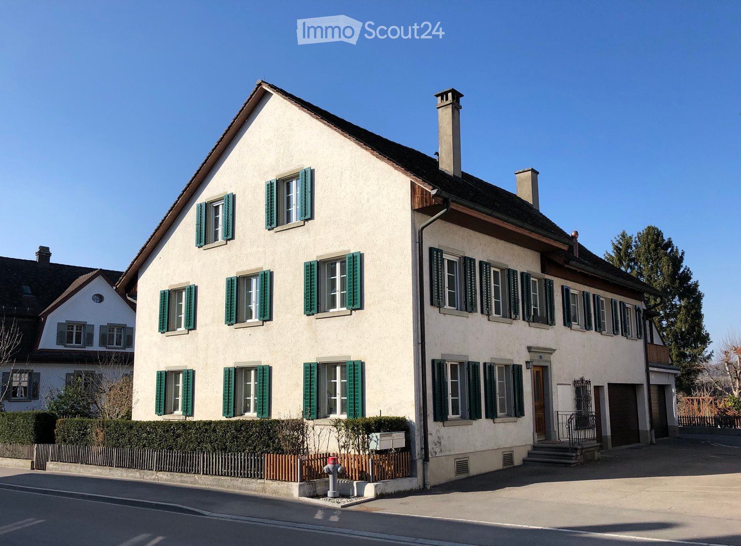 Two-story house with white walls, green shutters, and multiple windows. Features a chimney, a small garden with a fence, and a front staircase leading to the entrance.