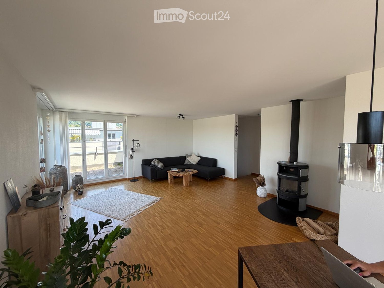 Modern living room with wooden flooring, large windows, a black sofa, a wooden coffee table, a fireplace, and decorative vases.
