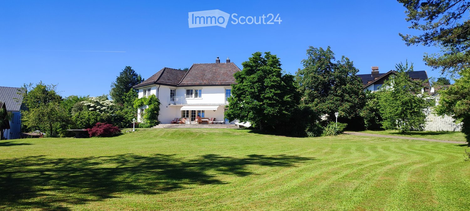 A two-story white house with a brown roof, surrounded by a well-maintained lawn and trees. The house features a balcony and outdoor seating area.